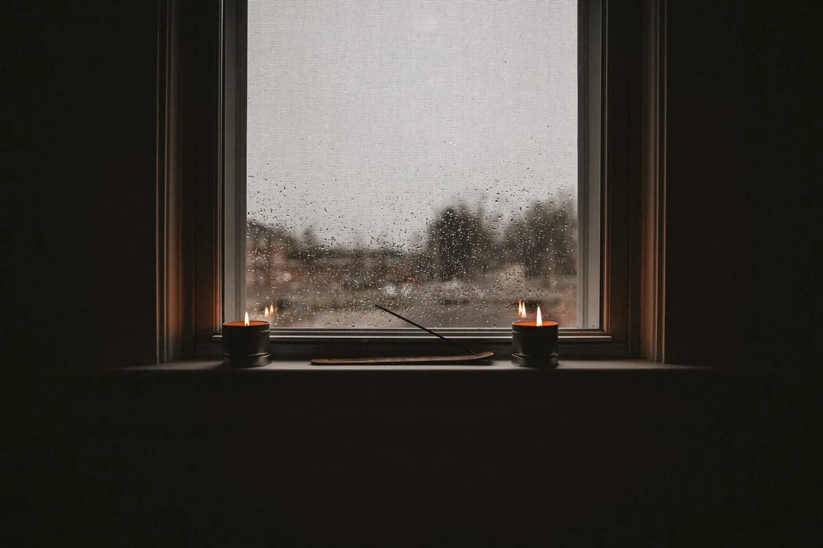 A dark room with two small candles on a windowsill and rain outside