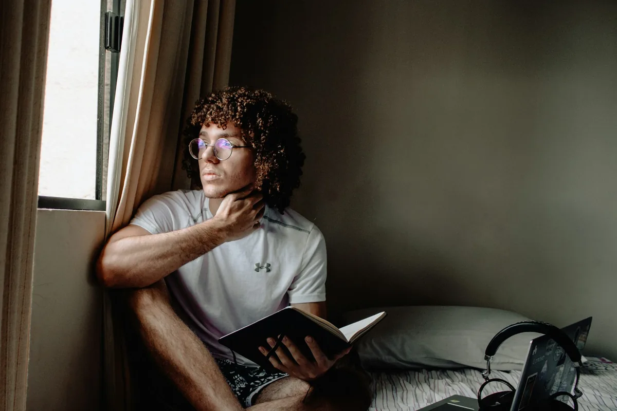 A young man sitting by a window with a book in his lap, looking outside